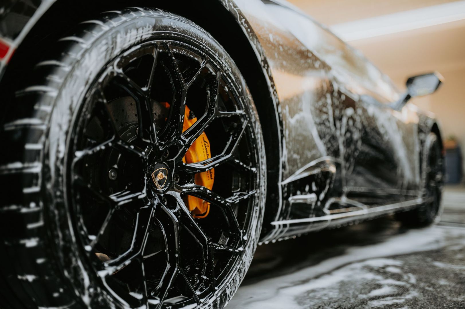Close-up of a black sports car wheel covered in soap during a car wash.